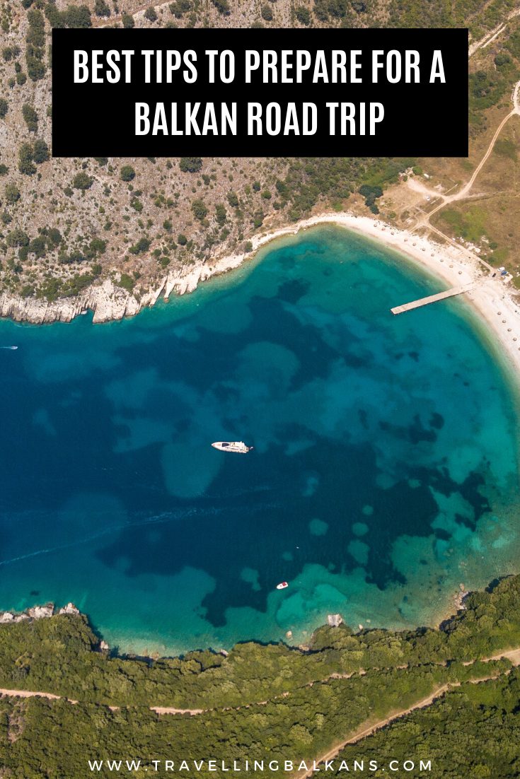 A bird's-eye view of a picturesque beach with turquoise water, a wooden pier, and boats dotting the sea. The image includes a bold overlay text reading “Best Tips to Prepare for a Balkan Road Trip” and the website URL “www.travellingbalkans.com.”