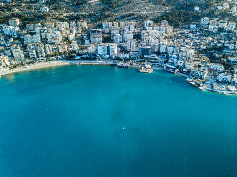 Aerial view of Sarandë, Albania, showing turquoise waters along a crescent beach lined with white high-rise buildings and rugged hills in the background.