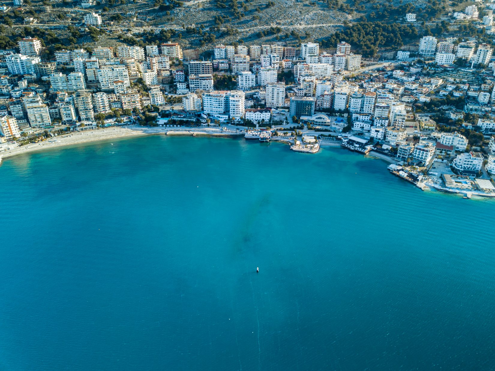 Aerial view of Sarandë, Albania, showing turquoise waters along a crescent beach lined with white high-rise buildings and rugged hills in the background.