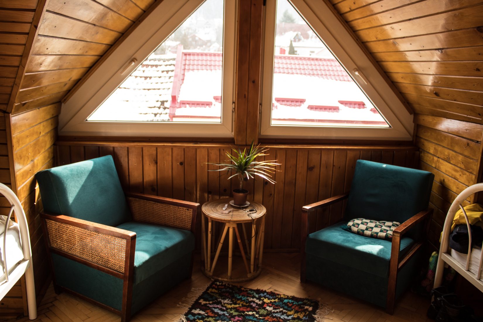 Cozy attic seating nook with two teal armchairs, a plant on a wooden table, and snowy rooftops seen through a triangle-shaped window.