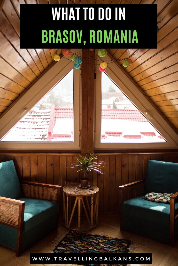 Cozy attic nook with two teal armchairs, a small wooden table, and views of snow-covered red rooftops through triangle windows.