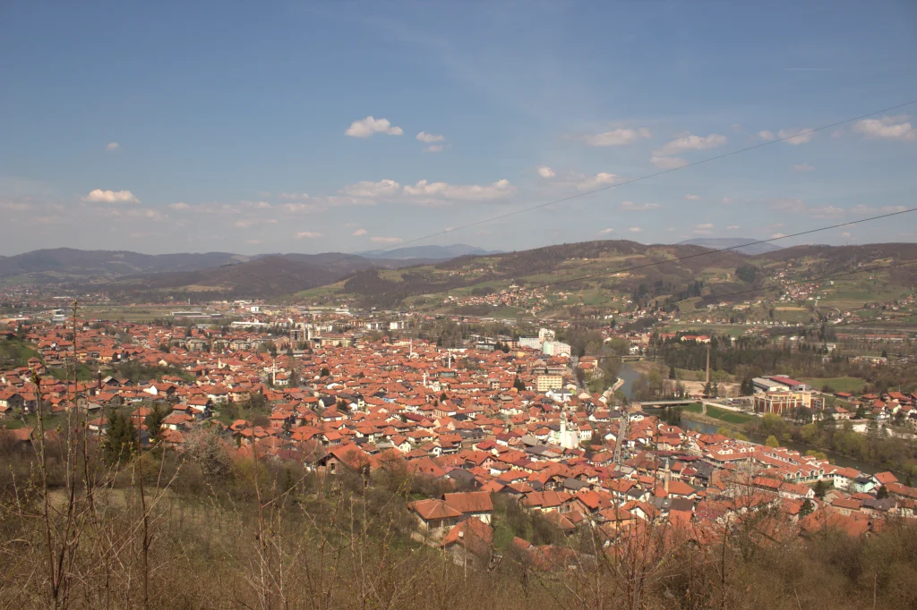 The image showcases a panoramic view of a town with a sea of red-tiled rooftops stretching across the landscape. The town is surrounded by rolling green hills and distant mountains, giving a sense of being nestled in a valley. The sky is bright and clear, with a few scattered clouds, providing a warm and inviting atmosphere. In the foreground, the town's buildings are densely packed, while beyond the town, the countryside is more spread out, with patches of farmland and smaller communities visible. This wide-angle view gives an expansive and peaceful look at a vibrant, rural setting.