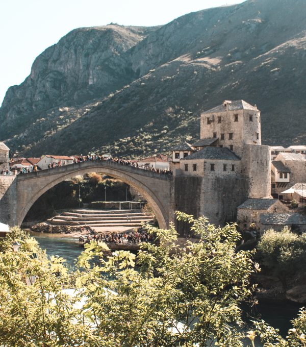 The image shows a picturesque view of a historic stone bridge, likely the famous Stari Most in Mostar, Bosnia and Herzegovina. The arched bridge spans a clear blue river, with people gathered along the top, taking in the scenery. On either side of the bridge, there are stone buildings with traditional rooftops, and the foreground is filled with green foliage. In the background, steep hills or mountains create a dramatic natural setting, with sunlight casting warm tones across the landscape. The image evokes a sense of history and beauty in this charming location.