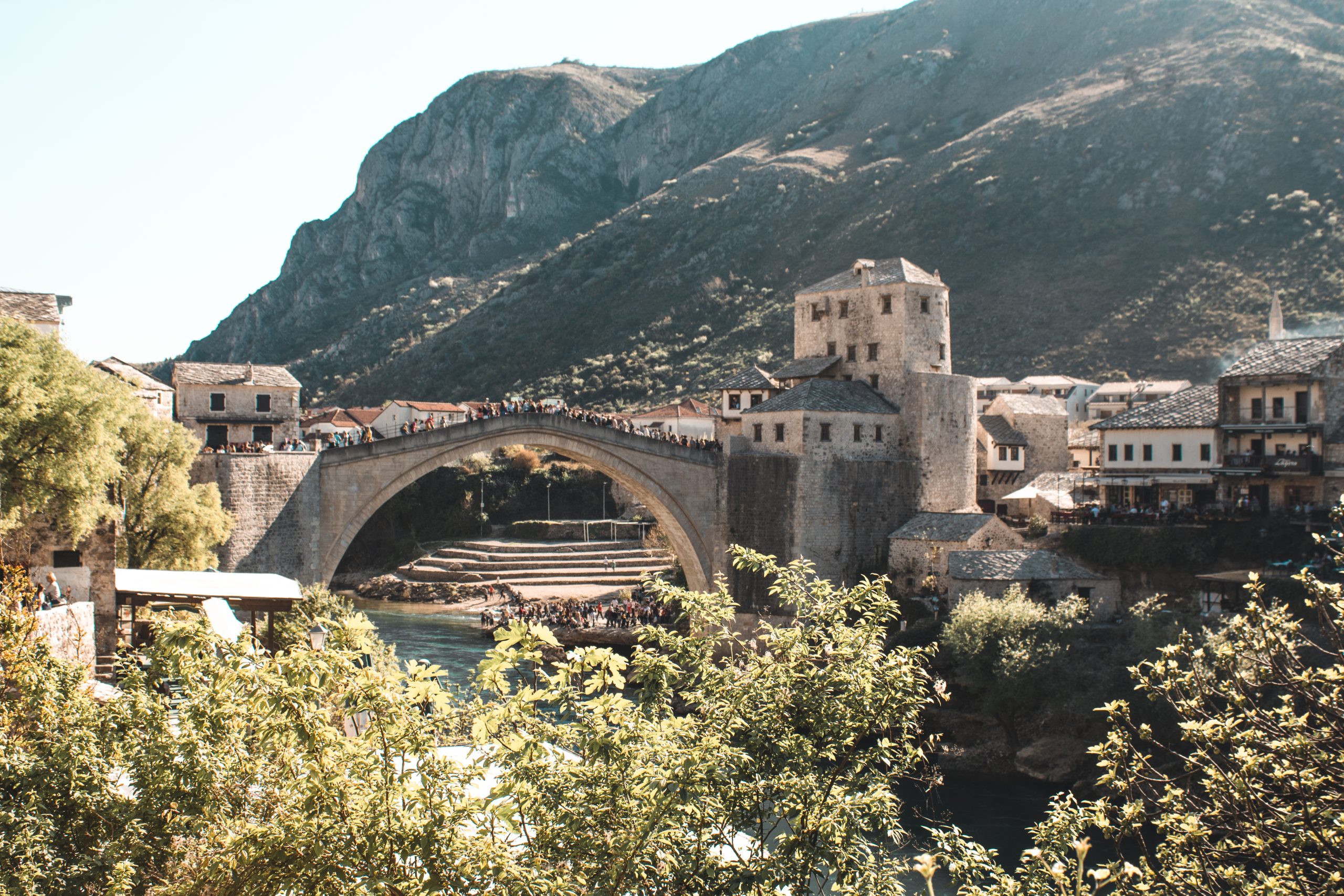 The image shows a picturesque view of a historic stone bridge, likely the famous Stari Most in Mostar, Bosnia and Herzegovina. The arched bridge spans a clear blue river, with people gathered along the top, taking in the scenery. On either side of the bridge, there are stone buildings with traditional rooftops, and the foreground is filled with green foliage. In the background, steep hills or mountains create a dramatic natural setting, with sunlight casting warm tones across the landscape. The image evokes a sense of history and beauty in this charming location.
