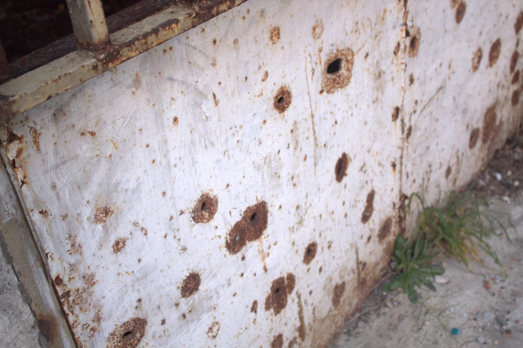The image shows a weathered metal surface, likely part of a door or wall, with multiple rusted holes and corrosion marks. The surface is white, but the rust has formed around and inside the holes, giving it a worn and aged appearance. The metal also shows signs of scratching and general wear. At the bottom right corner, there is some overgrown grass and debris.