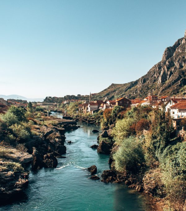 The image captures a stunning landscape of a river winding through a small town with traditional houses featuring red-tiled roofs. The river is a vibrant blue, cutting through rocky terrain and lush greenery on both sides. In the background, steep, rugged hills dominate the scene, creating a dramatic natural setting. The clear, bright sky adds a sense of calm and openness to the photo. The town appears peaceful, with the buildings nestled along the riverbank, blending harmoniously with the natural surroundings. The view suggests a charming, quiet locale surrounded by beautiful nature.