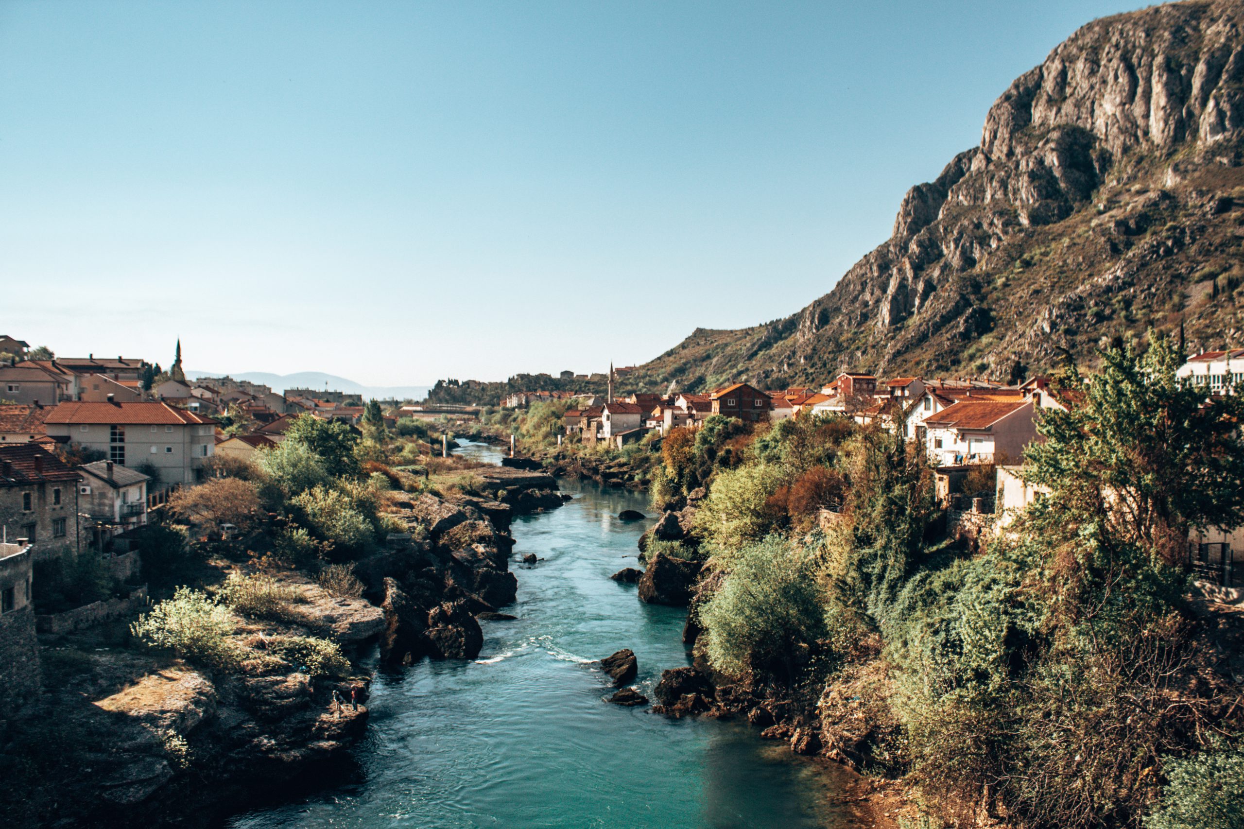 The image captures a stunning landscape of a river winding through a small town with traditional houses featuring red-tiled roofs. The river is a vibrant blue, cutting through rocky terrain and lush greenery on both sides. In the background, steep, rugged hills dominate the scene, creating a dramatic natural setting. The clear, bright sky adds a sense of calm and openness to the photo. The town appears peaceful, with the buildings nestled along the riverbank, blending harmoniously with the natural surroundings. The view suggests a charming, quiet locale surrounded by beautiful nature.